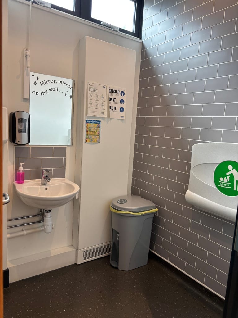 A gender neutral room with a wash basin, nappy bin, mirror and foldable changing table in. 