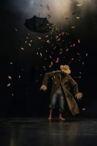 A scarecrow stands on a stage, with autumn leaves blowing around, and an umbrella suspended above them