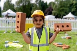 Girl wearing high visibility vest, hard hat and holding 2 toy bricks