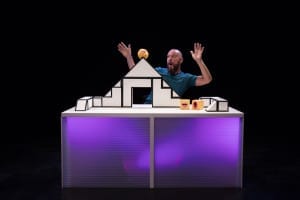 A man in a blue t shirt sits behind a model of a house in black and white on a purple table