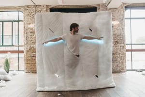 A man lays on a duvet with lights under his arms like wings in a bright studio