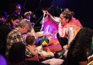 A boldly dressed actress holding up a prop to baby