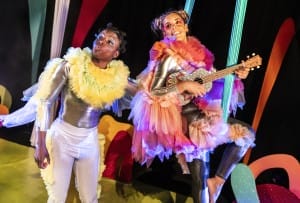 2 boldly dressed actresses on set, one holding a guitar