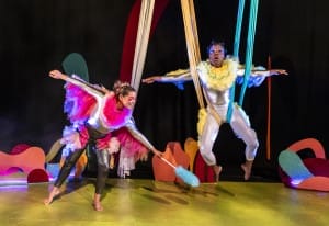 2 people hang from the ceiling in colourful silks, one has a feather duster and they're both pulling shocked expressions