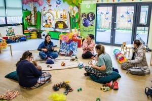 Lady with black hair sings with a guitar in a circle of parents and babies in a bright, colourful room