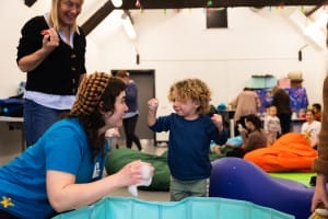 Facilitator Izzy wears a bright blue t shirt, and a woolly hat - playing in a paddling pool full of foam with a toddler
