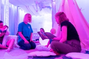 Karina, a facilitator wears a bright blue T shirt, and reads with a parent in a purple-lit sensory room